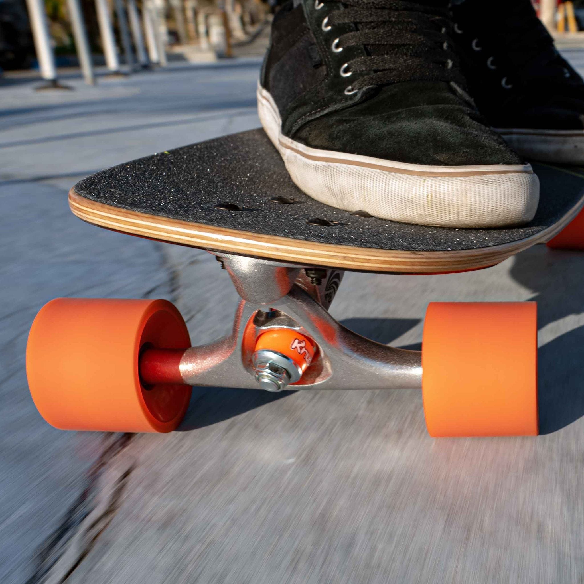 Skater's shoe on a longboard whose truck is fitted with orange (soft) Orangatang Knuckles bushings, orange wheels, motion blur