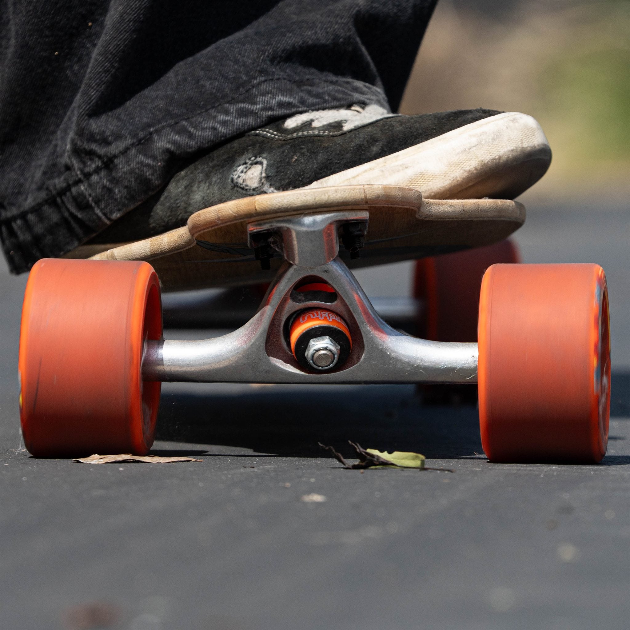 Low view of a skater's shoe on a longboard with orange (soft) Orangatang Nipples bushings in the truck and orange wheels