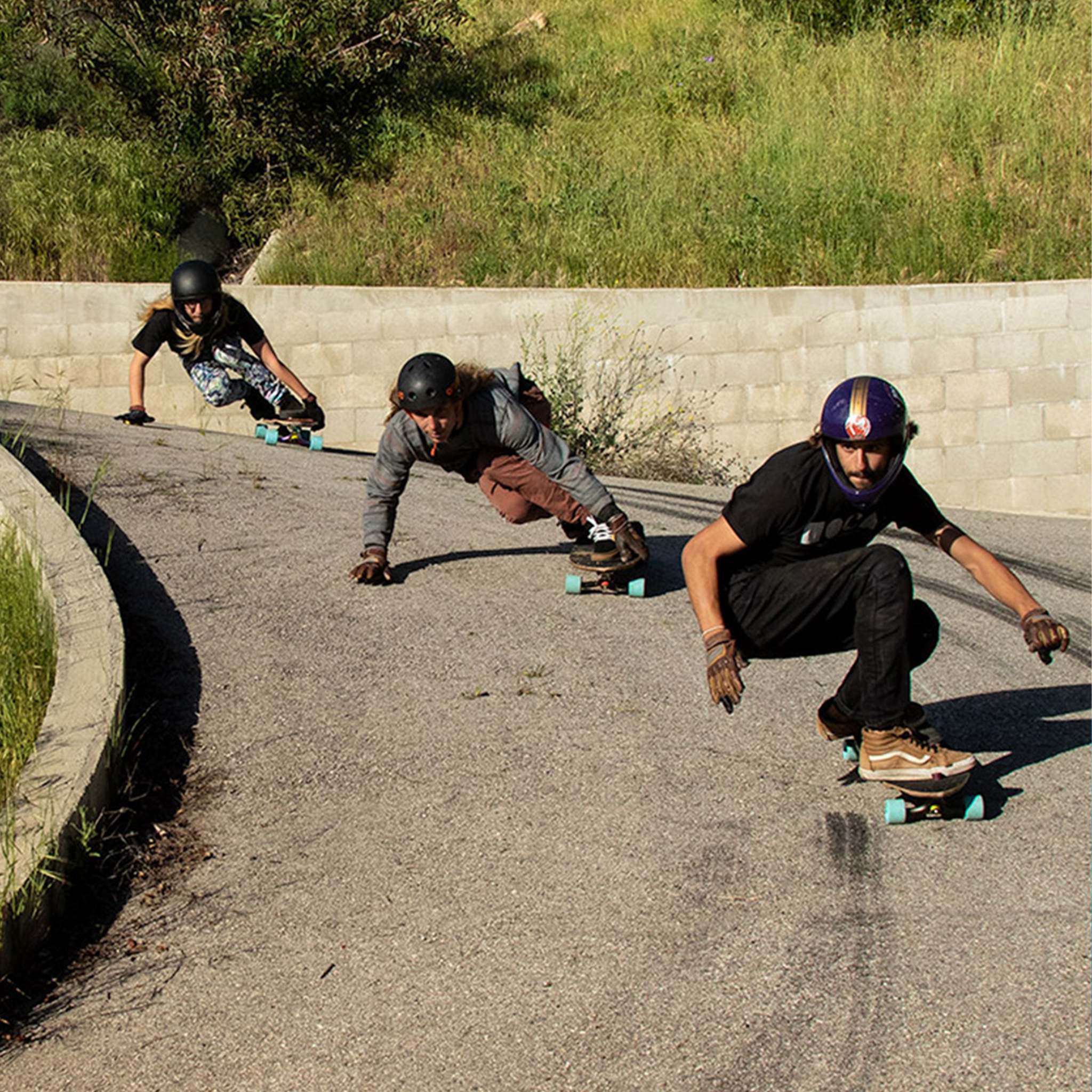 Three longboarders in helmets and slide gloves carving a paved downhill turn on Orangatang 4President 70mm wheels