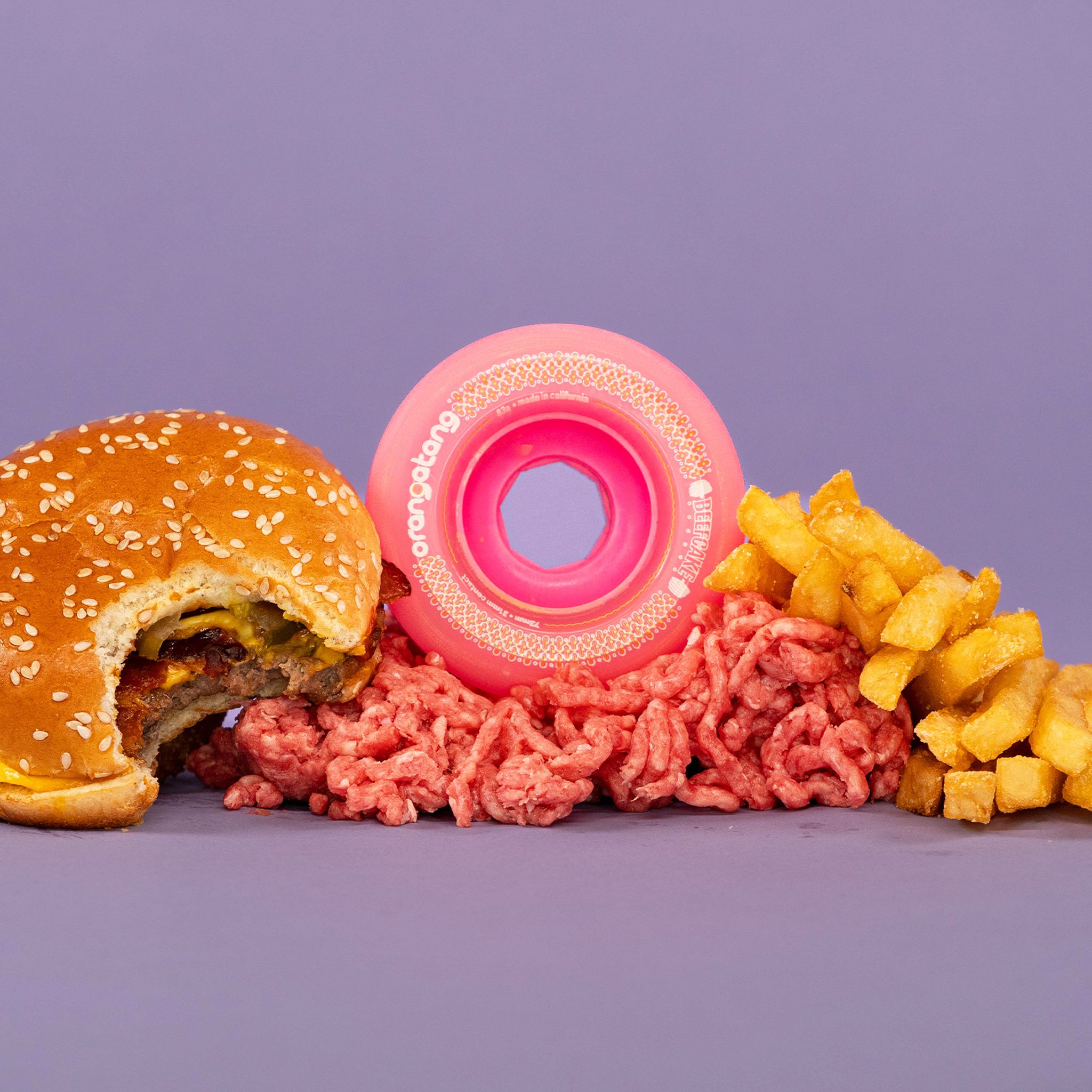 Pink Orangatang Beefcake 73mm wheel posed with a cheeseburger, raw ground beef, and fries on a purple background