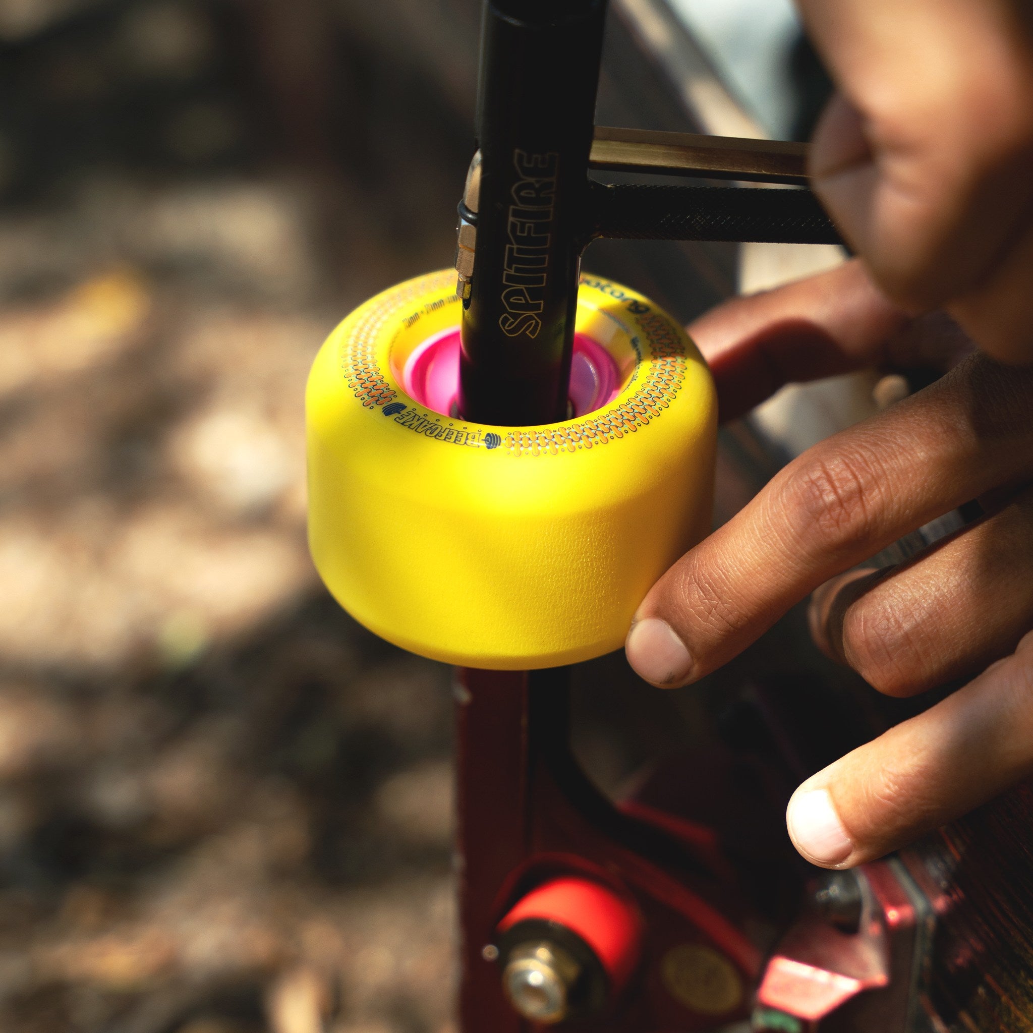 Hands using a skate tool to mount a yellow Orangatang Beefcake 73mm wheel with pink core onto a red skateboard truck