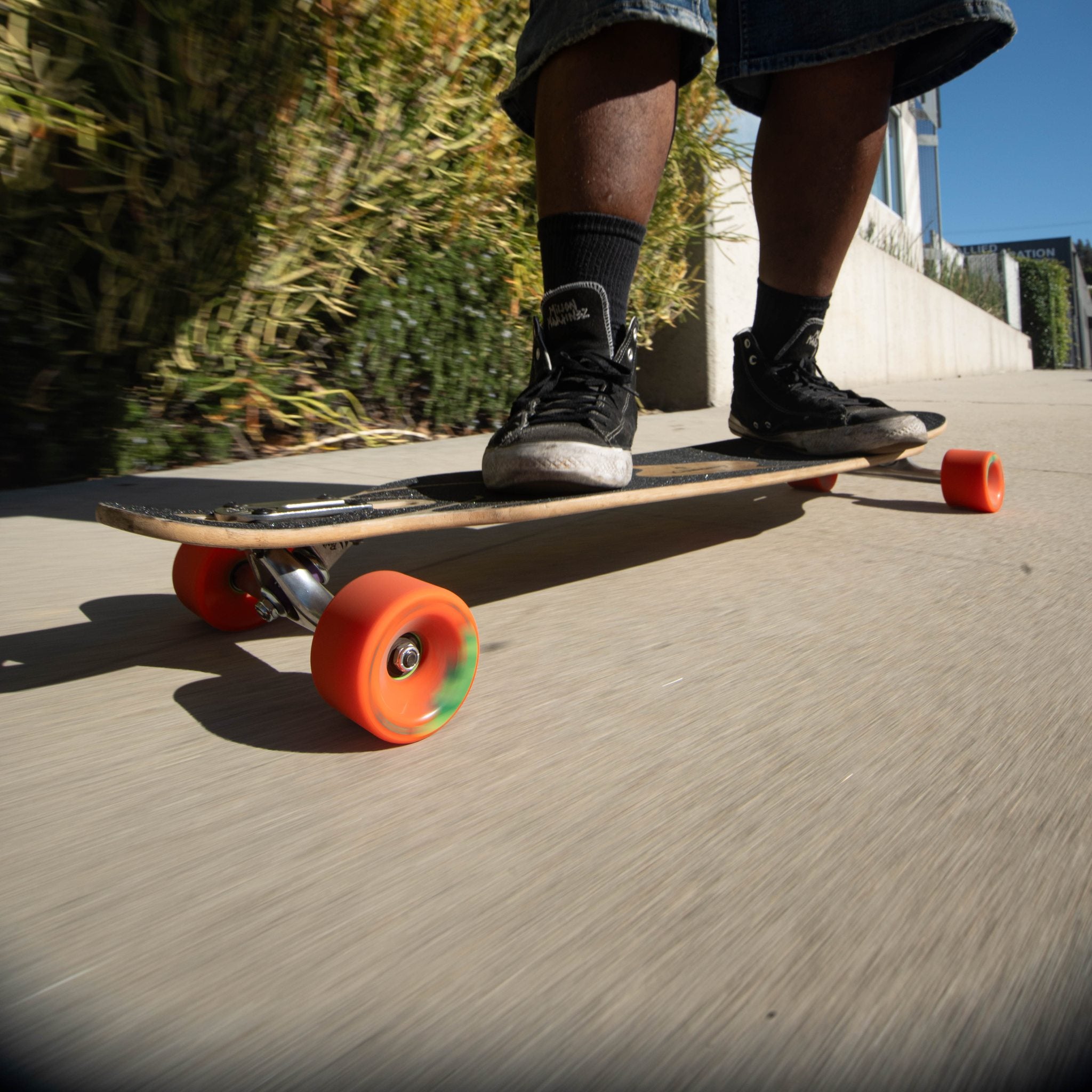 Close-up of a skater's feet on a longboard rolling on orange Orangatang Durian 75mm wheels on a sunny concrete path