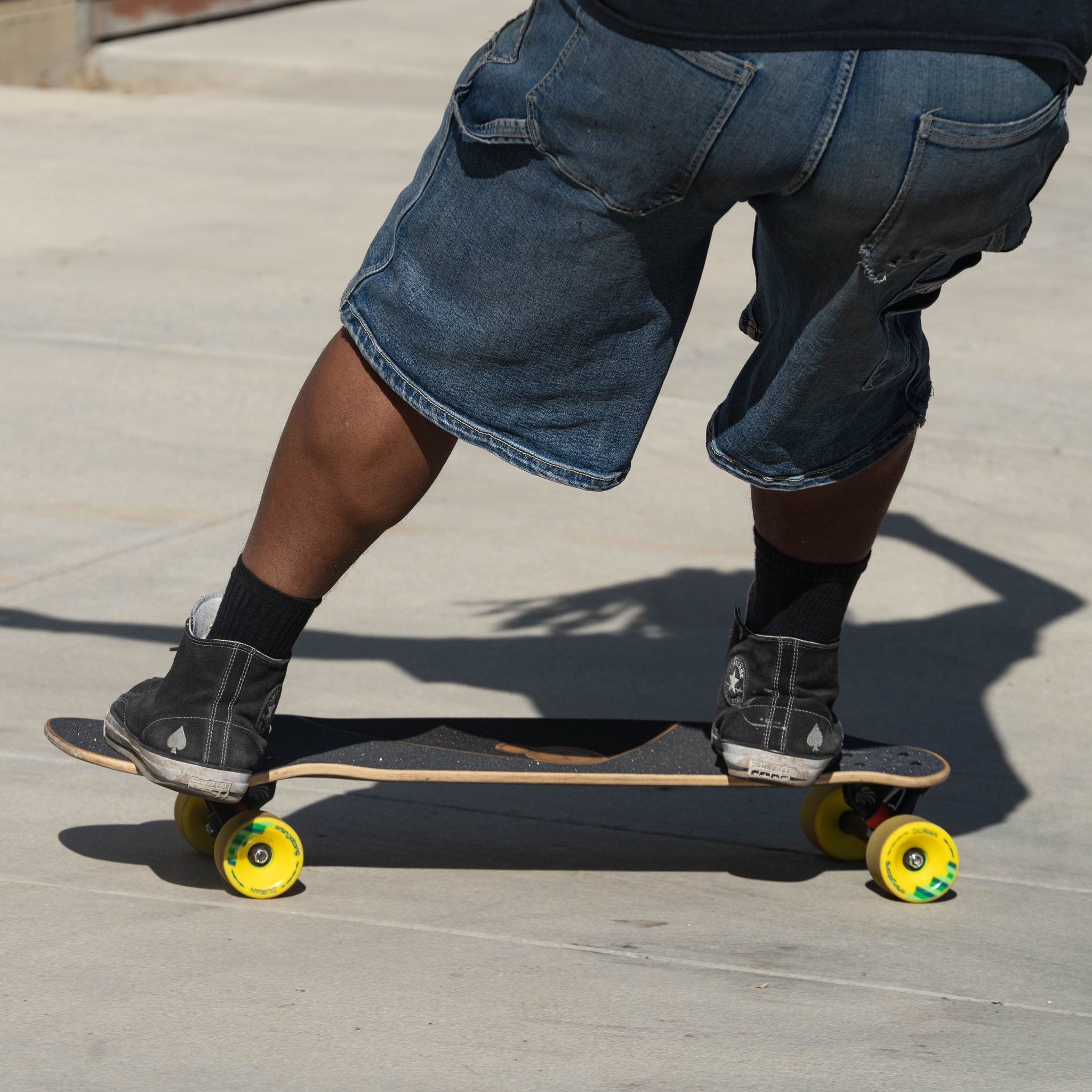 Skater in denim shorts and high-tops pushing a longboard fitted with yellow Orangatang Durian 75mm wheels on concrete
