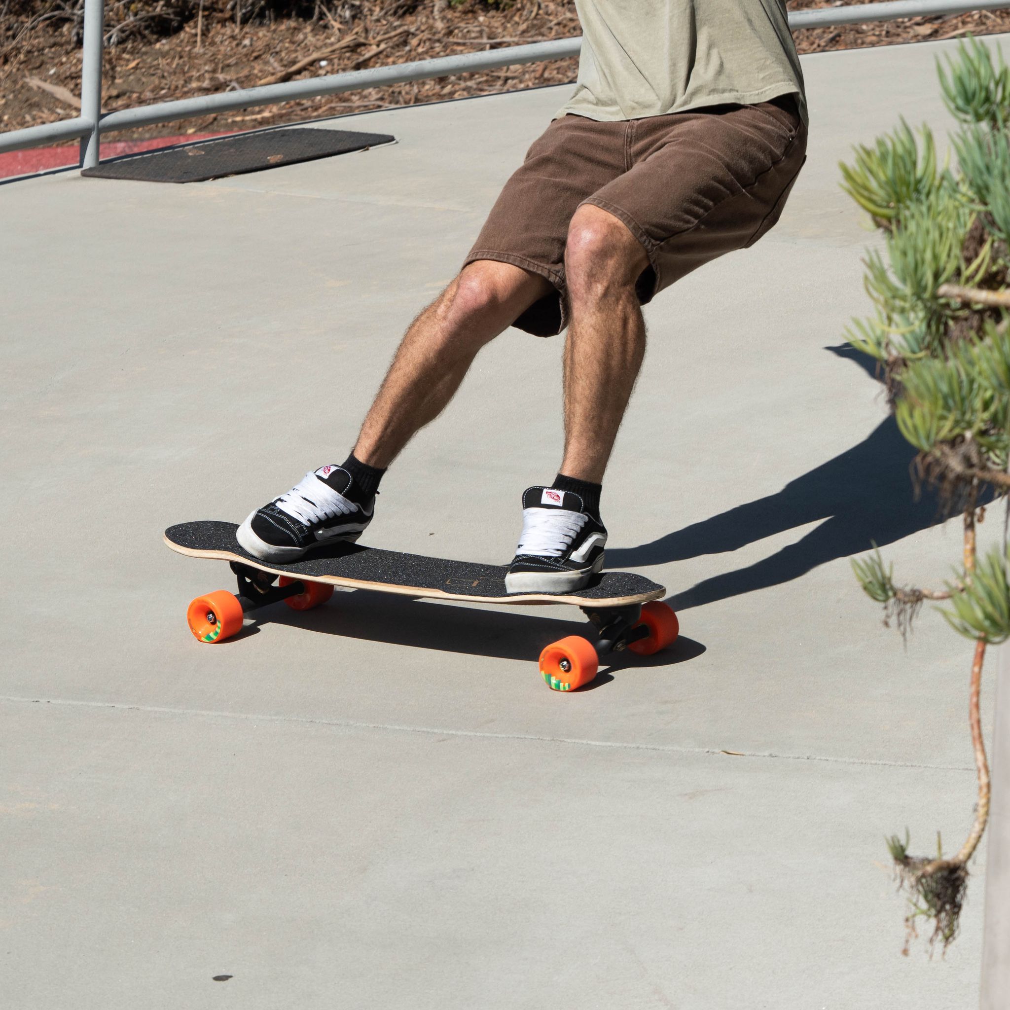 Skater in shorts carving a longboard with orange Orangatang Durian 75mm wheels on a smooth concrete plaza