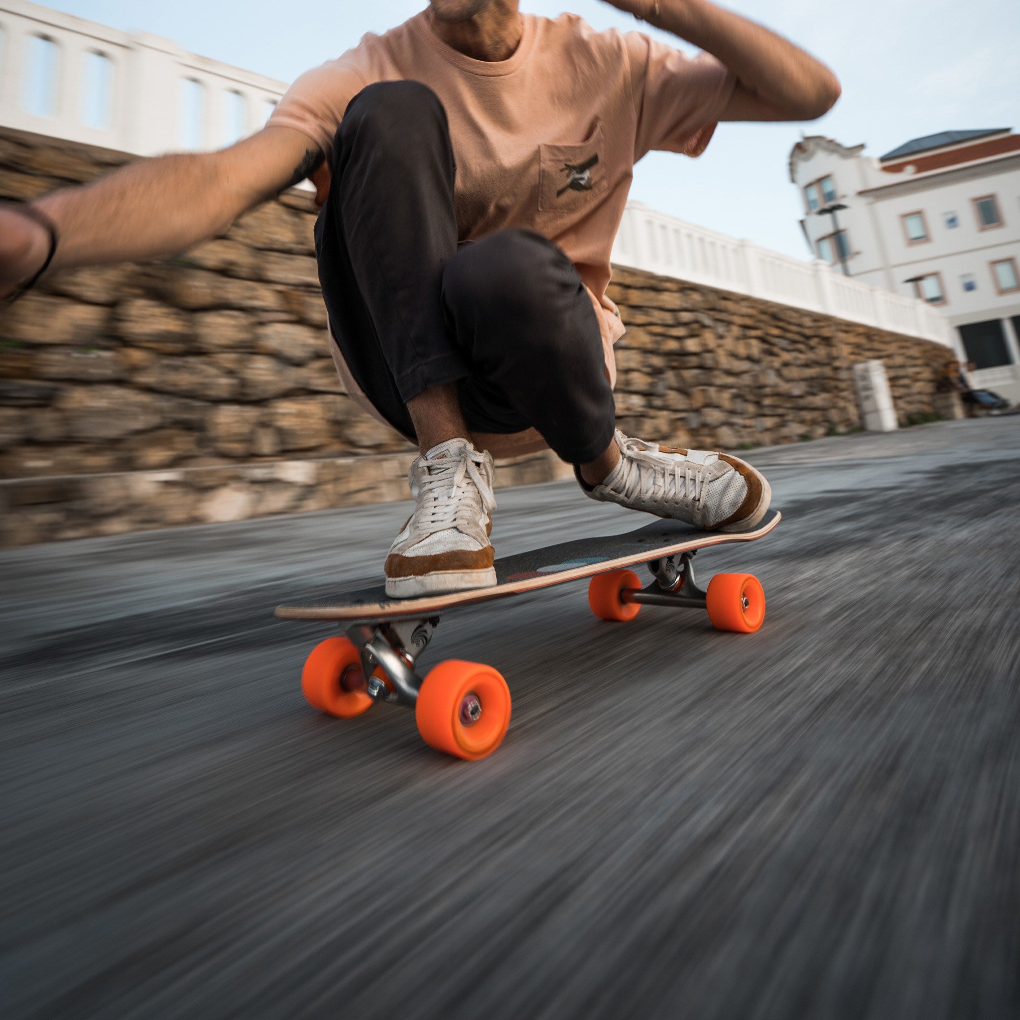 Skater crouched low on a cruiser board with orange Orangatang Fat Free 65mm wheels, speeding past a stone wall