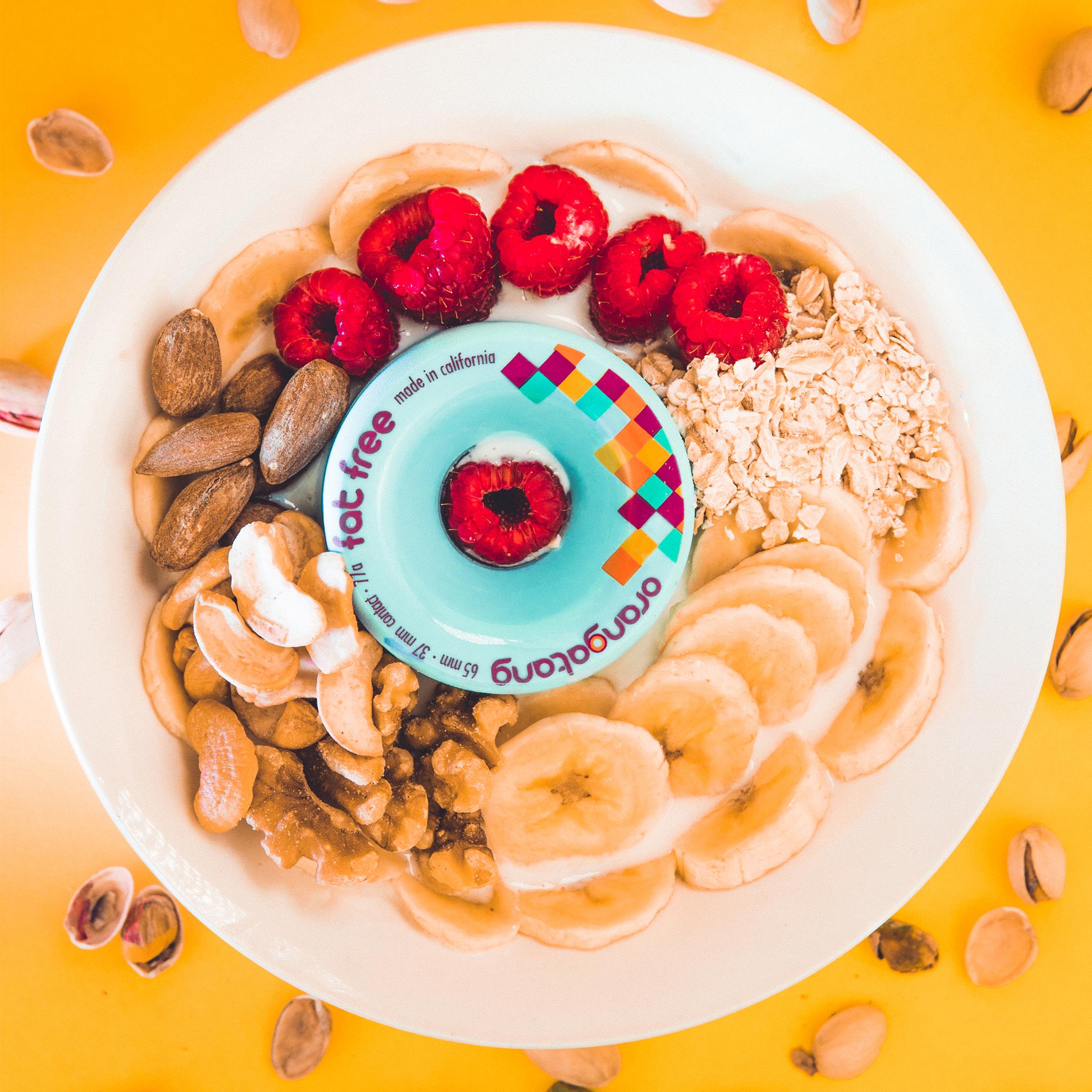 Blue Orangatang Fat Free 65mm wheel placed in a breakfast bowl with bananas, raspberries, oats, and nuts on a yellow background
