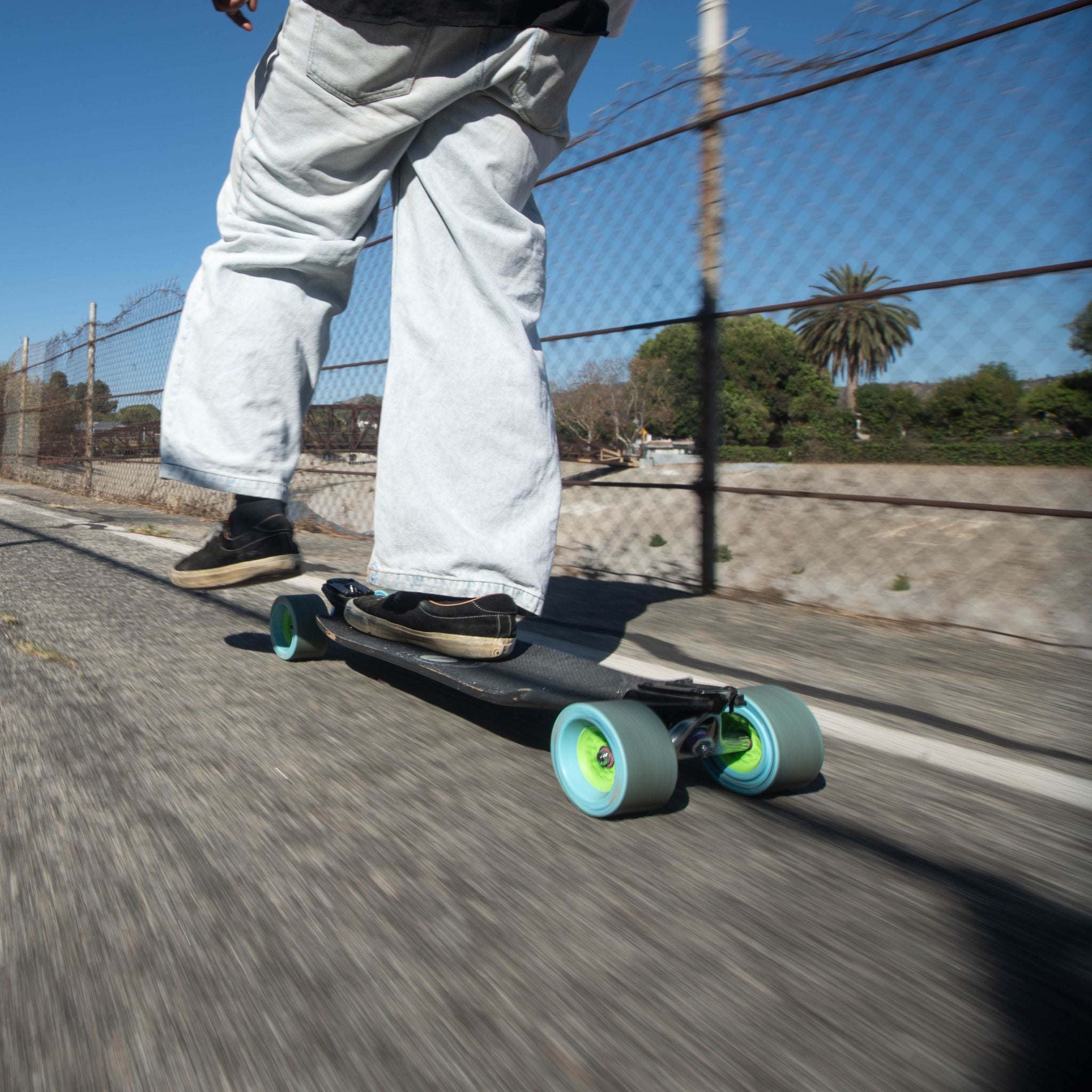 Rider in wide-leg jeans cruising a longboard on light blue Orangatang Dad Bod 105mm wheels along a fenced path