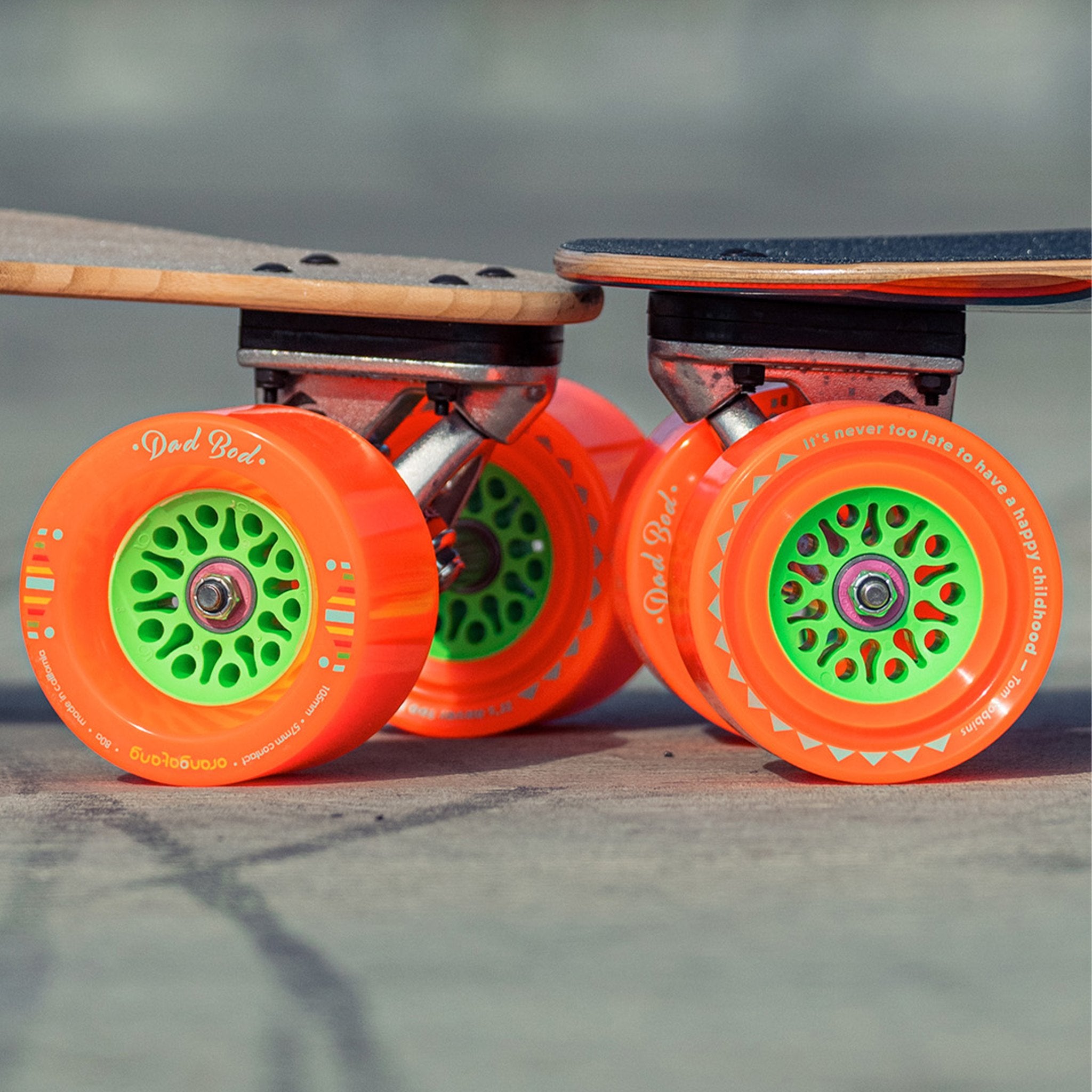 Close-up of orange Orangatang Dad Bod 105mm wheels with green cores mounted on the trucks of two longboards