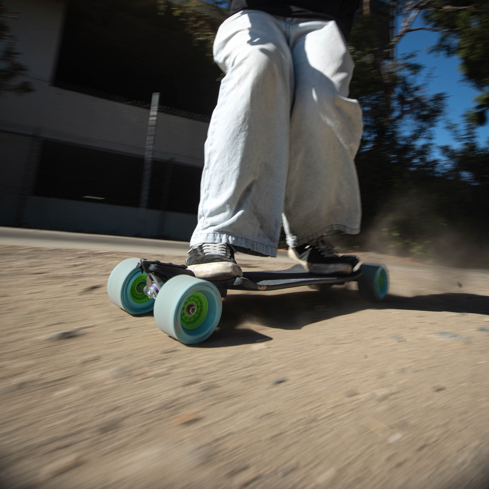 Rider in baggy jeans kicking up dust on a dirt path on a longboard with light blue Orangatang Dad Bod 105mm wheels