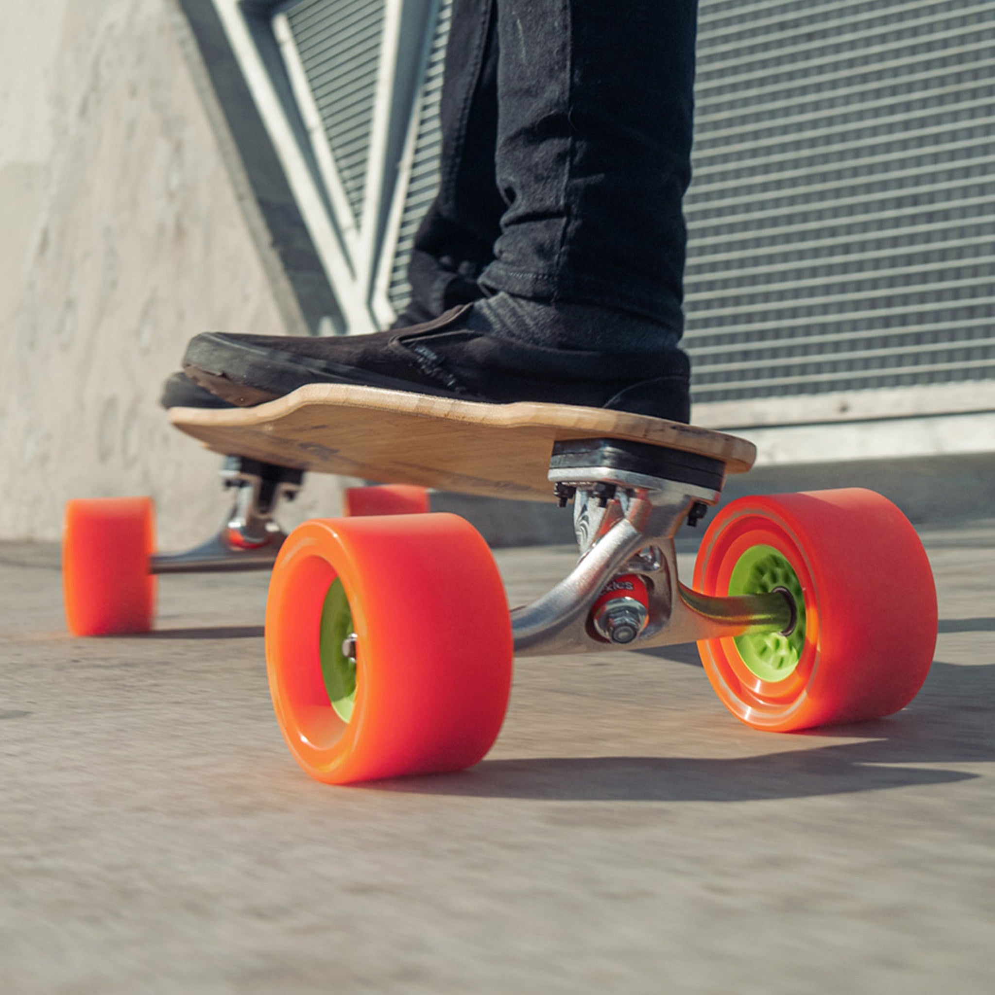 Ground-level view of orange Orangatang Dad Bod 105mm wheels with green cores under a longboard, rider's shoe on the deck