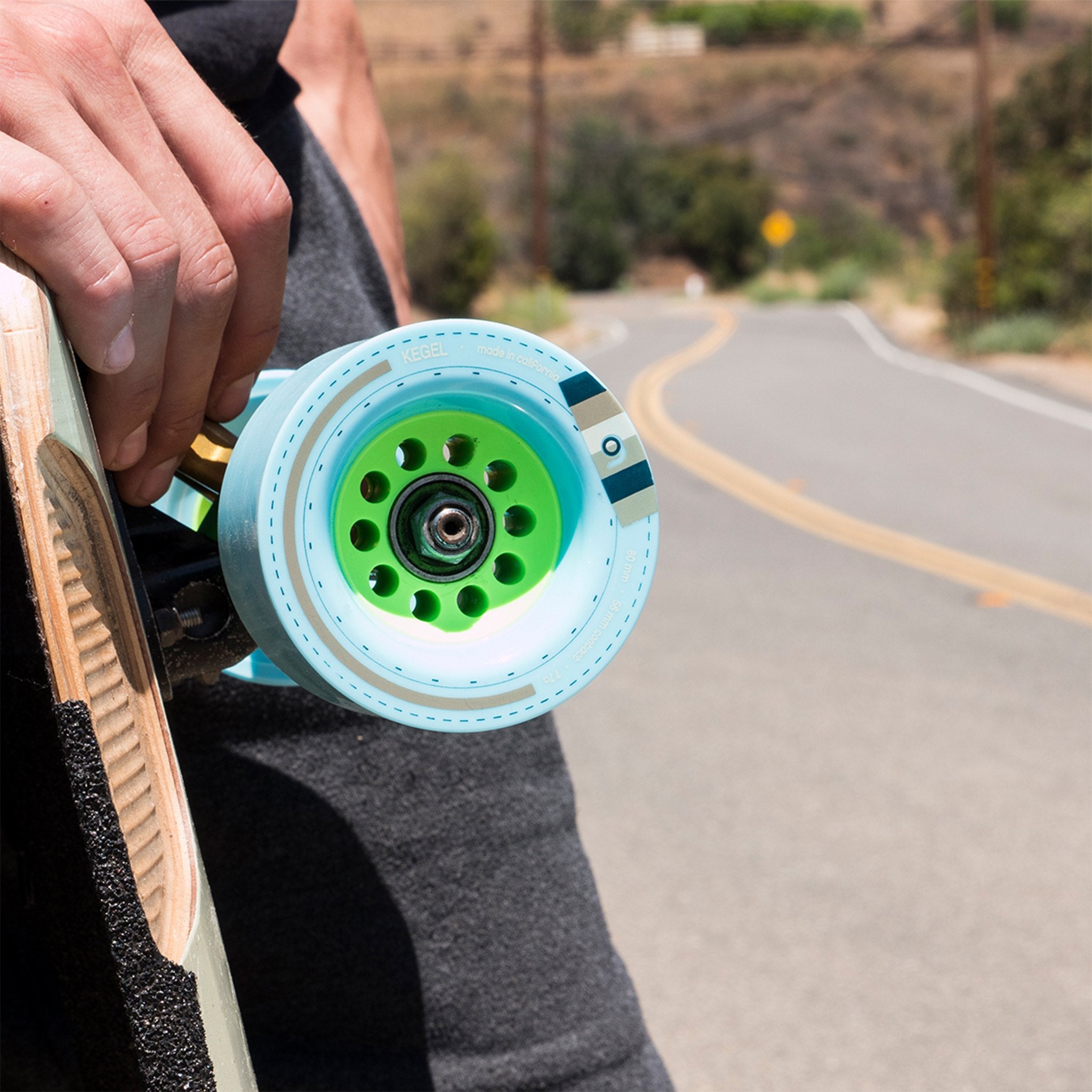 Skater carrying a longboard with light blue Orangatang Kegel 80mm wheels and green cores on a winding rural road