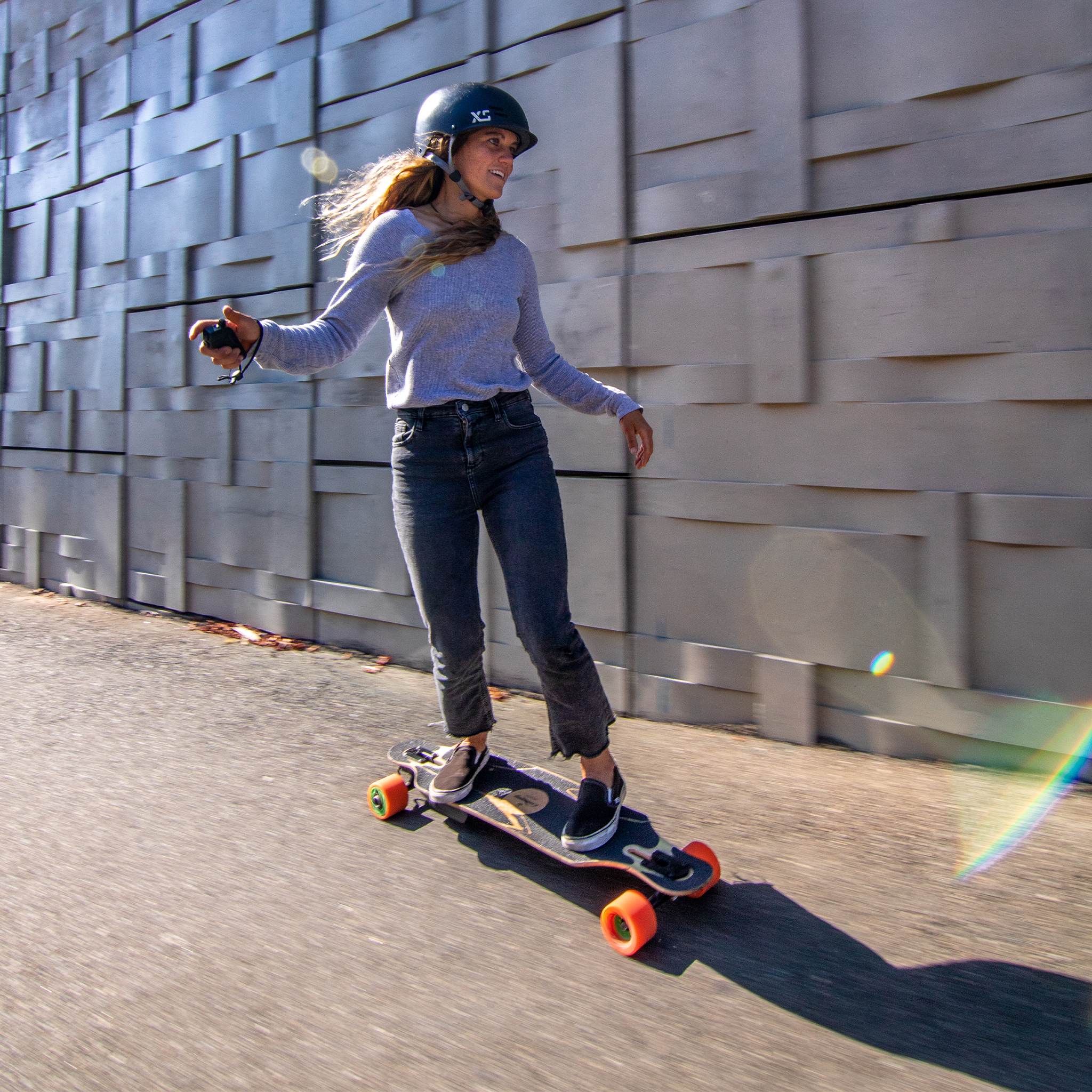 Rider in a helmet cruising a longboard with orange Orangatang Kegel 80mm wheels past a textured concrete wall