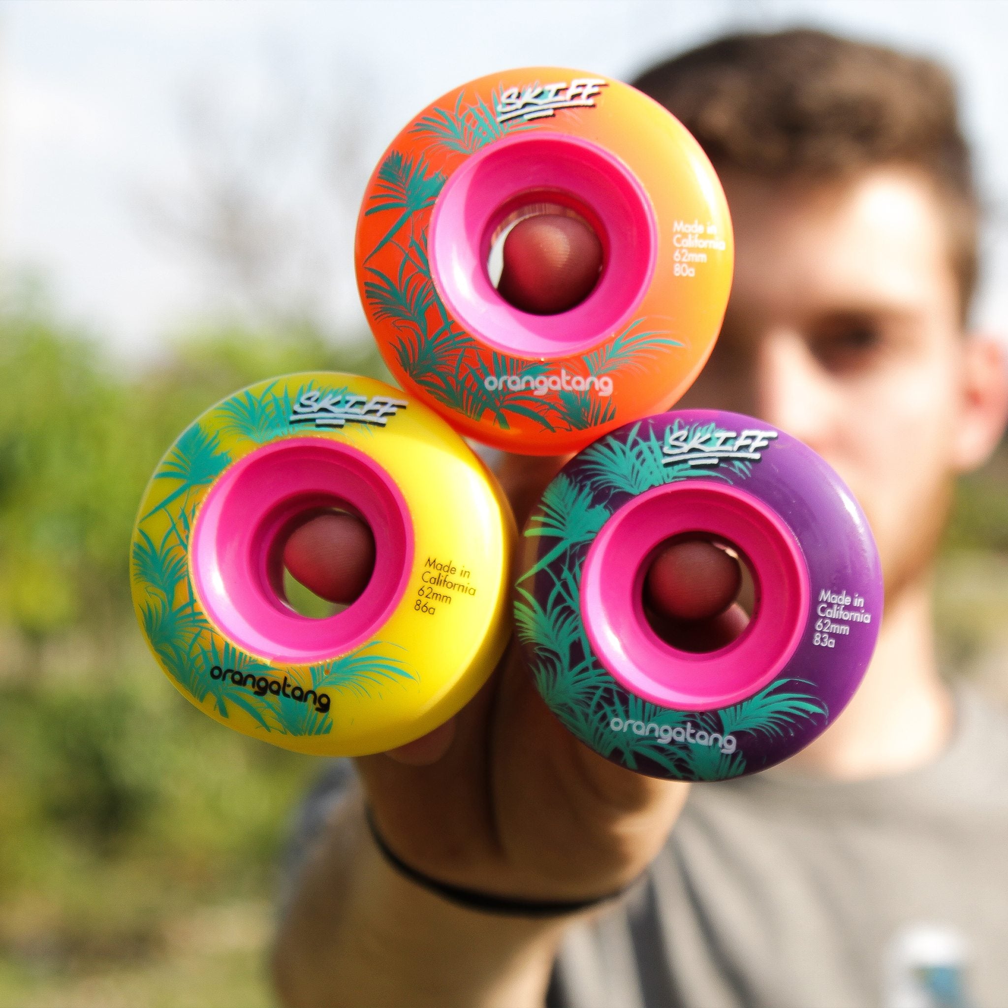 Skater holding up three Orangatang Skiff 62mm wheels in orange, yellow, and purple with pink cores and palm leaf graphics